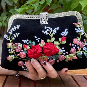 Op de afbeelding: Een zwarte fluwelen clutch met een zilveren sluiting en bloemenborduursel. Het borduursel toont rode, roze en witte bloemen met groene bladeren.