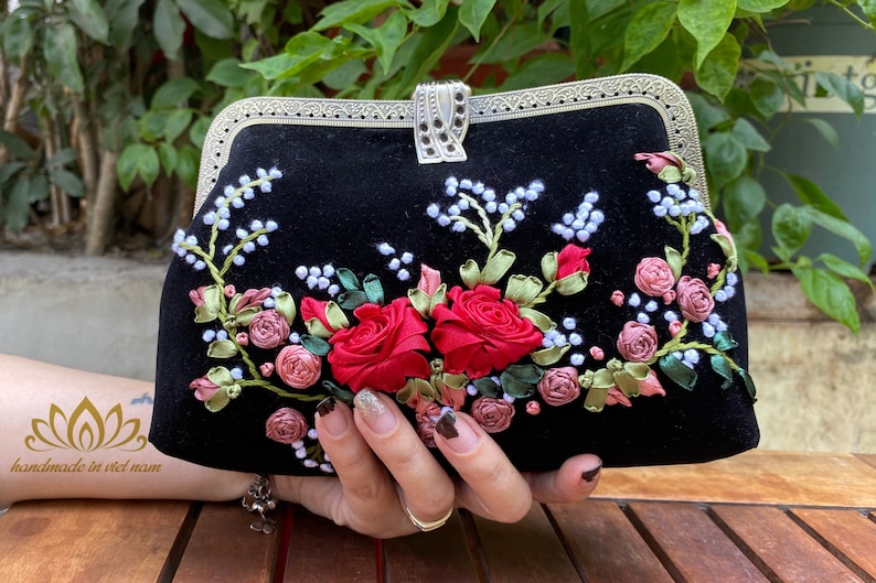 Op de afbeelding: Een zwarte fluwelen clutch met een zilveren sluiting en bloemenborduursel. Het borduursel toont rode, roze en witte bloemen met groene bladeren.