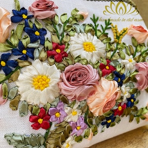 May include: A white fabric pouch with a floral design embroidered with colorful ribbon. The design features roses, daisies, and other flowers in shades of pink, white, blue, red, and yellow. The pouch has the text "handmade in viet nam" embroidered on the top.