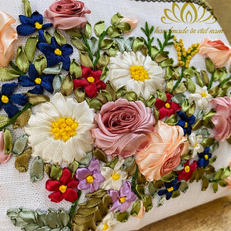 May include: A white fabric pouch with a floral design embroidered with colorful ribbon. The design features roses, daisies, and other flowers in shades of pink, white, blue, red, and yellow. The pouch has the text "handmade in viet nam" embroidered on the top.