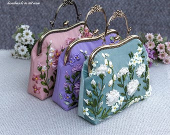 May include: Three structured handbags in pink, lavender, and teal, each featuring a metallic kiss-lock frame and handle. The fabric surfaces are decorated with intricate ribbon embroidery depicting various floral stems, leaves, and white roses.