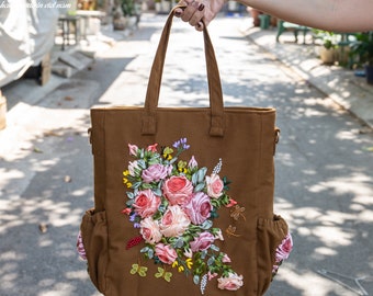 May include: A brown canvas tote bag with a floral design embroidered in pink, white, and green ribbon. The bag has two side pockets and a top handle.