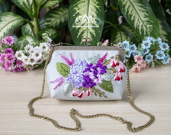 Può includere: Una pochette in satin bianco con un ricamo floreale con fiori di nastro viola, rosa e verde. La borsa ha una tracolla a catena dorata.