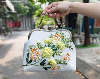 Può includere: Una pochette bianca con un motivo floreale ricamato con nastro giallo, arancione e verde. La pochette ha una chiusura in metallo e un manico superiore.