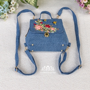 Pode incluir: Uma pequena mochila de ganga azul com um design de bordado floral. O bordado apresenta flores cor-de-rosa, vermelhas e verdes com abelhas amarelas. A mochila tem alças ajustáveis e um fecho dourado.