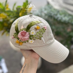 May include: A beige baseball cap decorated with embroidered flowers, leaves, and insects. The floral design features a pink rose, white daisies, and yellow insects. The cap has a curved peak and a stitched pattern.