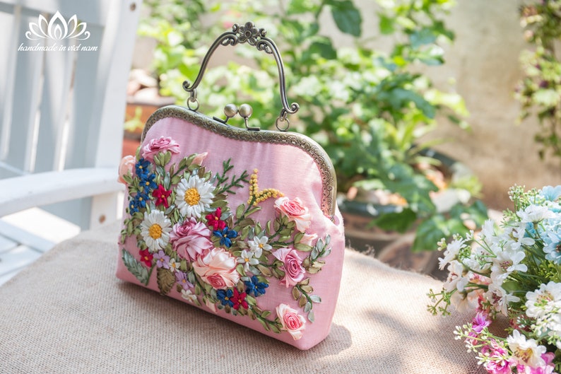 May include: A pink satin clutch bag with a floral embroidery design. The embroidery features pink, white, blue, and green ribbon flowers.