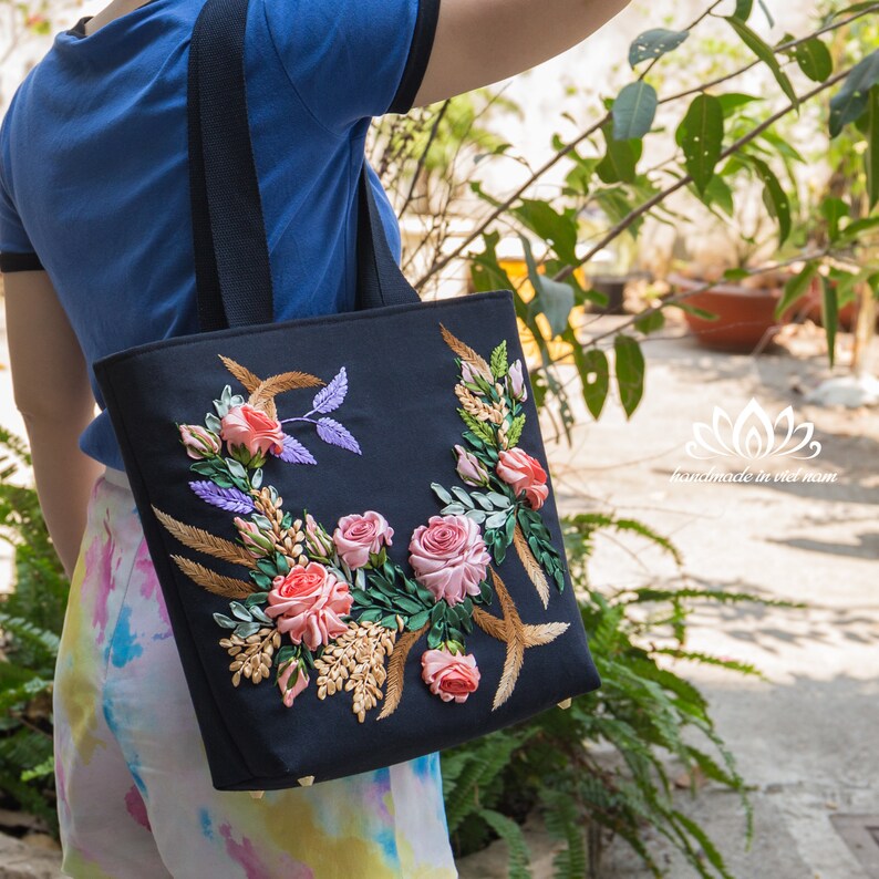 K&ouml;nnte beinhalten: Eine schwarze Tragetasche mit einem floralen Design, das mit rosa, pfirsichfarbenem und gr&uuml;nem Band bestickt ist. Die Tasche hat schwarze Henkel und goldfarbene Beschl&auml;ge.