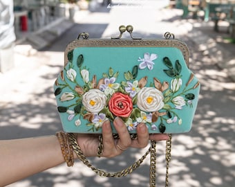 以下が含まれることがあります： 花柄の刺繍が施されたターコイズブルーのベルベット製クラッチバッグ。刺繍には白、ピンク、緑のリボンで作られた花が施されています。バッグにはゴールドのチェーンストラップが付いています。