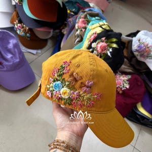 Puede incluir: Una gorra de béisbol de color amarillo mostaza con un diseño de bordado floral. El bordado presenta flores rosas, azules y amarillas, hojas verdes y detalles de mariposas. La gorra tiene una visera curvada y un cierre de velcro.