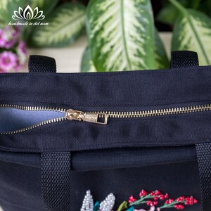 K&ouml;nnte beinhalten: Eine schwarze Canvas-Tragetasche mit einem goldenen Rei&szlig;verschluss und einem floralen Stickmuster. Die Tasche hat zwei schwarze Henkel.