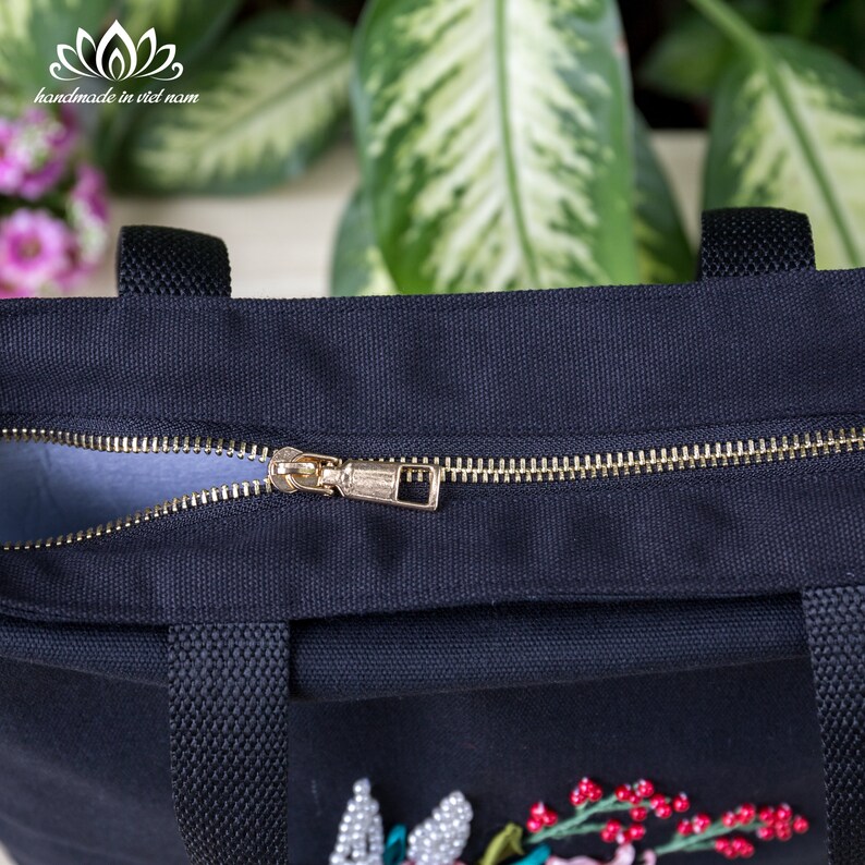 K&ouml;nnte beinhalten: Eine schwarze Canvas-Tragetasche mit einem goldenen Rei&szlig;verschluss und einem floralen Stickmuster. Die Tasche hat zwei schwarze Henkel.