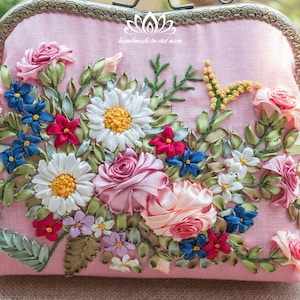 May include: A pink fabric clutch purse with a floral embroidery design. The embroidery features pink, white, blue, and green ribbon flowers.