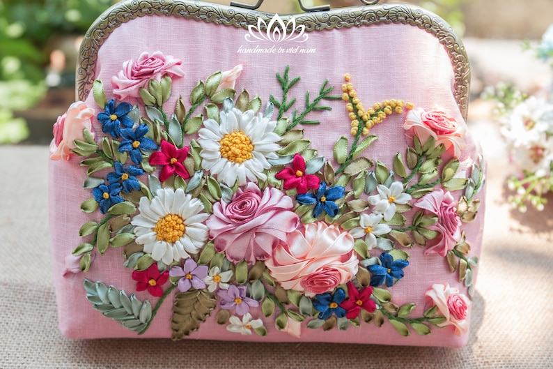 May include: A pink fabric clutch purse with a floral embroidery design. The embroidery features pink, white, blue, and green ribbon flowers.