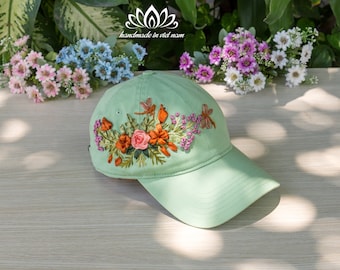 May include: A light green baseball cap with a floral embroidery design featuring pink and orange flowers, green leaves, and dragonfly accents. The embroidery is handmade in Vietnam.