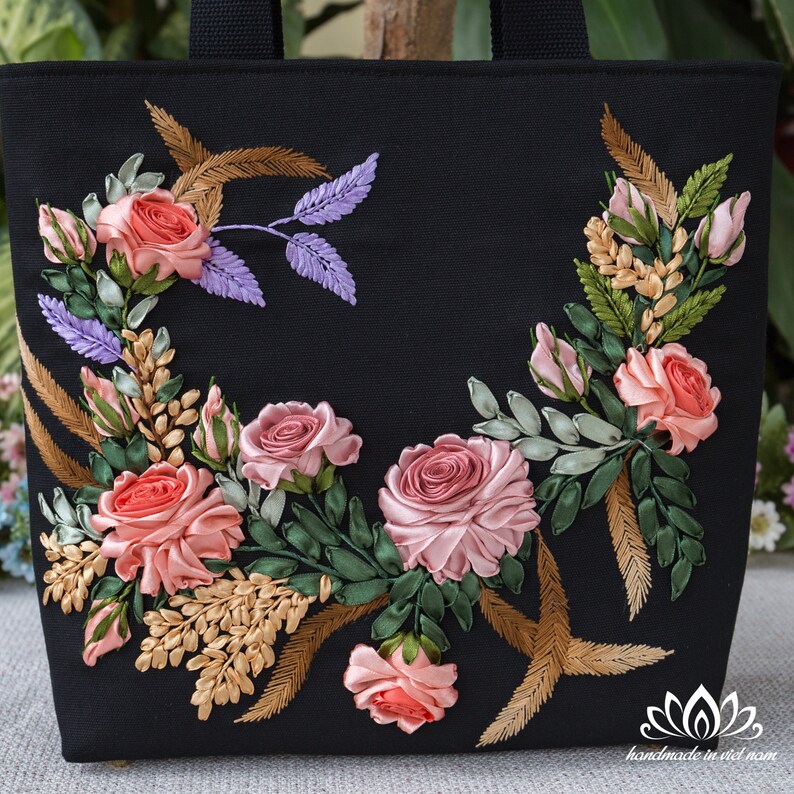 K&ouml;nnte beinhalten: Schwarze Canvas-Tragetasche mit einem floralen Design, das mit rosa, gr&uuml;nem und goldenem Band bestickt ist. Das Design zeigt Rosen, Bl&auml;tter und Weizen&auml;hren.