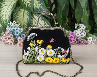 May include: A black velvet handbag with a floral embroidery design. The bag has a metal clasp closure and a chain strap. The embroidery features yellow, white, and purple flowers, as well as green leaves and a few red butterflies.