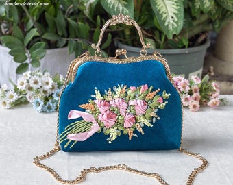 May include: A teal velvet handbag featuring a gold-tone metal frame, a top handle, and a detachable gold chain strap. The front is decorated with a three-dimensional floral arrangement made of pink and green ribbon embroidery and a pink ribbon.