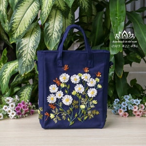May include: A navy blue tote bag with a floral design embroidered with white, yellow, and green ribbon. The bag has two top handles and a detachable shoulder strap. The text "handmade in viet nam 0905.422.456" is printed on the bag.