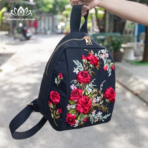 Puede incluir: Mochila negra con diseño floral. La mochila presenta un bordado de rosas rojas vibrantes con hojas verdes y flores blancas. Tiene una cremallera dorada y el texto "handmade in viet nam".
