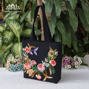 K&ouml;nnte beinhalten: Schwarze Tragetasche mit einem floralen Design, das mit rosa, pfirsichfarbenem und gr&uuml;nem Band bestickt ist. Die Tasche hat schwarze Henkel und ein schwarzes Futter.