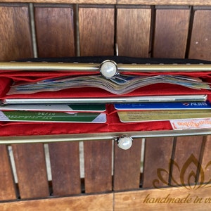 Op de afbeelding: Een zwarte leren portemonnee met een gouden metalen frame en een rode voering. De portemonnee is open en toont meerdere creditcards en contant geld van binnen.