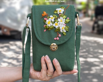 May include: A green canvas crossbody bag with a flap closure and a gold clasp. The bag features a floral embroidery design with white and green ribbon flowers. The bag has a long adjustable strap.