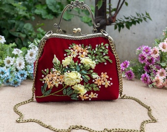 Puede incluir: Un bolso de mano de terciopelo rojo con un diseño de bordado floral. El bolso tiene una correa de cadena dorada y un cierre de metal.