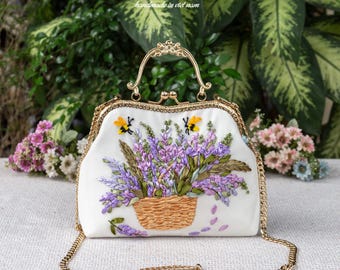 May include: A white handbag featuring a gold metal frame and a gold chain strap. The front is decorated with a raised ribbon embroidery of a woven basket filled with purple lavender flowers and green leaves, accented by two small yellow bees.