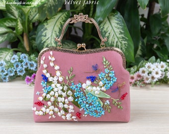 Può includere: Una borsa a mano in velluto rosa con chiusura e manico dorati. La borsa è decorata con fiori ricamati in tonalità di bianco, blu e verde.