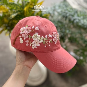 Puede incluir: Gorra de béisbol de color coral con bordados florales. La gorra presenta una visera curva y flores bordadas en tonos rosas y blancos. El texto "handmade in viet nam" es visible en la parte superior de la gorra.