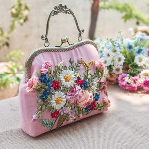 May include: A pink fabric clutch bag with a floral embroidery design. The embroidery features a variety of flowers in shades of pink, white, blue, and green. The bag has a metal clasp and a top handle.