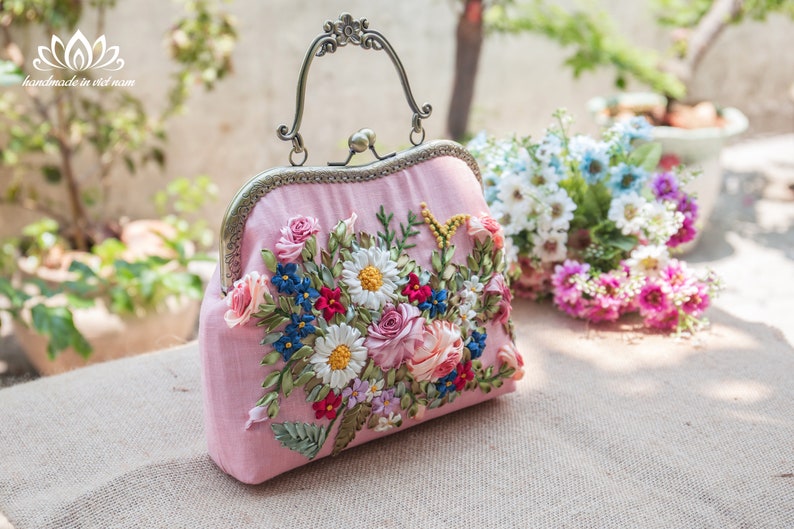 May include: A pink fabric clutch bag with a floral embroidery design. The embroidery features a variety of flowers in shades of pink, white, blue, and green. The bag has a metal clasp and a top handle.