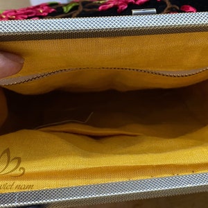 May include: Inside view of a black velvet clutch bag with a yellow fabric lining. The bag has a zipper closure and a small pocket inside. The bag is embroidered with pink flowers and has the text "handmade in viet nam" printed on the lining.