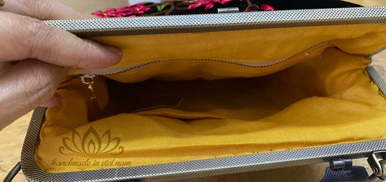May include: Inside view of a black velvet clutch bag with a yellow fabric lining. The bag has a zipper closure and a small pocket inside. The bag is embroidered with pink flowers and has the text "handmade in viet nam" printed on the lining.