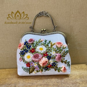 May include: A white fabric clutch purse with a floral embroidery design. The embroidery features pink, red, blue, and green silk ribbon flowers. The purse has a brass clasp and a top handle.