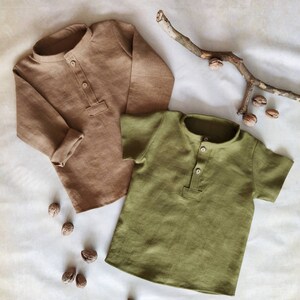 May include: Two linen shirts, one long-sleeved in a tan color and the other short-sleeved in olive green. Both shirts have a mandarin collar and a buttoned placket. Walnuts and a small branch are scattered around the shirts.