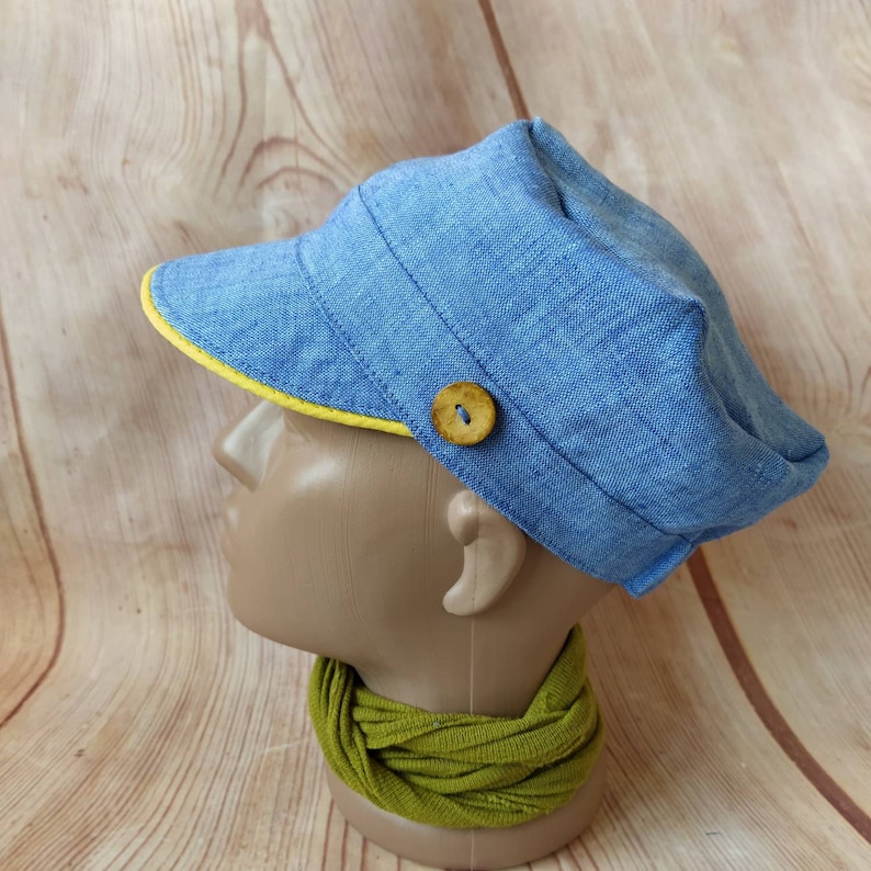 Pu&ograve; includere: Un berretto di jeans blu con un bordo giallo e un bottone di legno. Il berretto &egrave; su una testa di manichino.