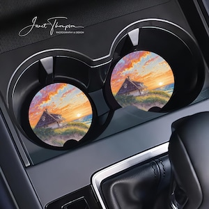 May include: Two car coasters with a sunset scene featuring a house on a grassy dune. The sky is filled with orange, pink, and yellow hues. The coasters are in a car's cup holders. The text "Janet Thompson PHOTOGRAPHY & DESIGN" is in the upper left.