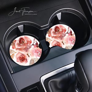 May include: Two car coasters with a watercolor floral design featuring red and pink roses and brown leaves on a white background. The coasters are placed in a car's cup holders.