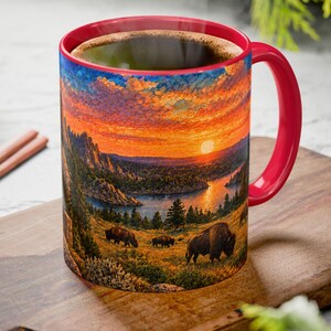 May include: A ceramic mug with a bright red handle and interior, featuring a wrap-around graphic of a sunset landscape with mountains, a lake, and three bison grazing in the foreground. The mug is filled with a dark liquid, likely coffee or tea.