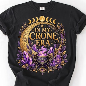 May include: A black t-shirt featuring a graphic print with the text IN MY CRONE ERA in gold lettering. The design includes a large golden crescent moon, a central cauldron with purple smoke, purple crystals, and a small glass bottle.