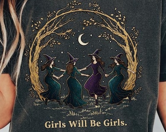 May include: Dark gray t-shirt featuring a circular illustration of witches dancing under a crescent moon and stars, framed by golden tree branches. The text "Girls Will Be Girls" is printed below the image. The shirt is worn by a person.