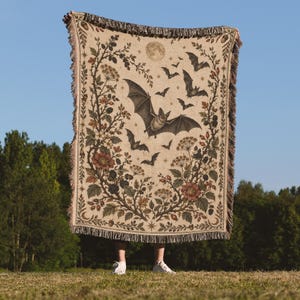 May include: A woven throw blanket featuring a cream-coloured background with a central illustration of a large bat surrounded by smaller bats, a full moon, and stars. The design is framed by intricate botanical vines with flowers and berries.