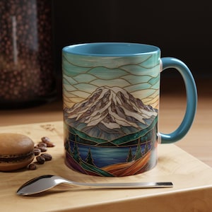 May include: A ceramic coffee mug with a blue handle and interior. The mug features a stained-glass style design of a mountain range, lake, and trees. The color palette includes blues, greens, browns, and yellows. A spoon and a macaron are on a wooden surface.