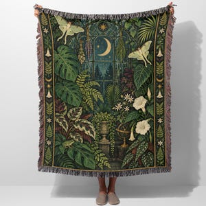 May include: A woven throw blanket featuring a dark green botanical scene with a large arched window, a crescent moon, two pale green moths, various tropical plants, ferns, white trumpet flowers, a small frog, and a decorative fringe border.