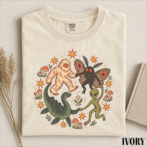 Può includere: Una t-shirt color avorio con etichetta Comfort Colors sul collo e una stampa grafica circolare raffigurante uno yeti beige, una falena nera, un dinosauro verde e un alieno verde che si tengono per mano, circondati da stelle arancioni e funghi rossi.