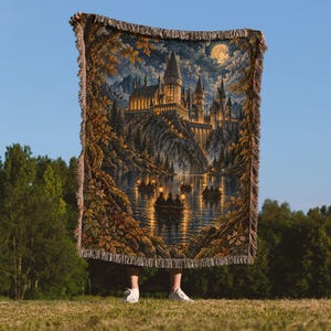 May include: A woven tapestry throw blanket featuring a detailed night scene of a large castle perched on a cliff above a lake. Small boats with glowing lanterns float on the water under a full moon. The blanket has a fringed edge and warm tones.