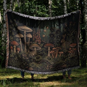 May include: A woven throw blanket featuring a dark forest scene with various mushrooms, a toad, a rabbit, and botanical elements like ferns and flowers. The blanket has a fringed edge and a detailed, multi-colored graphic design on a black background.