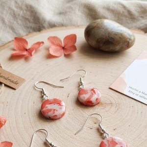 May include: Two pairs of drop earrings featuring round, polished charms with a marbled pattern of coral-orange and white. Each charm is attached to a silver-toned metal fishhook ear wire. The earrings are displayed on a light-colored wooden surface.